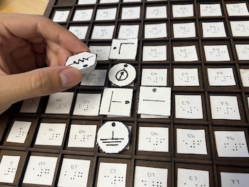 A hand places a round token with a squiggly line on a grid board with square tiles, each labeled with Braille and printed coordinates.