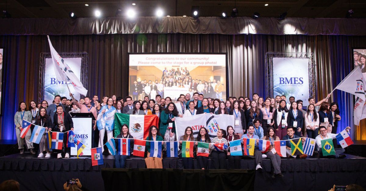 A large group of people stands on a stage holding various international flags at a BMES event, with a congratulatory message displayed on a screen in the background.
